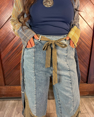 A person wearing light-wash denim patchwork shorts with brown jute trim and a matching jute tie belt, styled with a navy top and a yellow plaid shirt against a wooden paneled background.