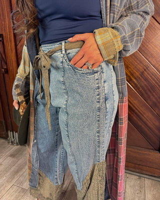 A person wearing a pair of wide-leg light-wash denim jeans with patchwork paneling and a brown drawstring waist, styled with a navy top, a gray plaid shirt, and a red plaid cardigan in front of a wooden door.