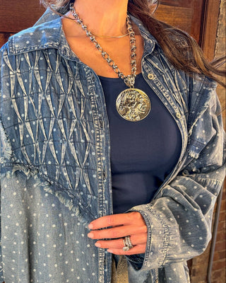 A woman wearing a light-wash blue denim shirt jacket with white embroidered dots and a crisscross pattern on the chest, layered over a navy blue top and light-wash jeans.
