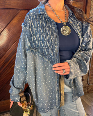 A woman wearing a light-wash blue denim shirt jacket with white embroidered dots and a crisscross pattern on the chest, layered over a navy blue top and light-wash jeans.