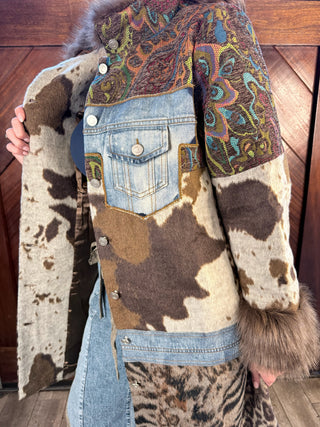A patchwork-style jacket with mixed materials including faux fur, cowhide, denim, and paisley, shown on a person in front of a wooden wall.