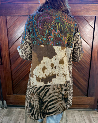 A person viewed from behind wearing a long patchwork coat with leopard print, cowhide, zebra print, and a colorful paisley pattern on the upper back, standing in front of a wooden door.