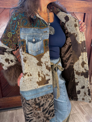 A person wearing a patchwork-style western-inspired coat with faux fur trim, denim pocket details, brown and cream cow-print panels, and leopard-print sections, paired with a navy top and light blue jeans in front of a wooden door.