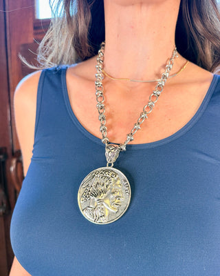 A close-up of a woman wearing a large silver-toned coin pendant necklace over a blue tank top.