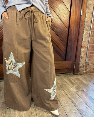 A person wearing wide-leg brown pants with embroidered star patches on the legs, paired with a light blue-gray top, standing in front of a wooden door and brick wall.