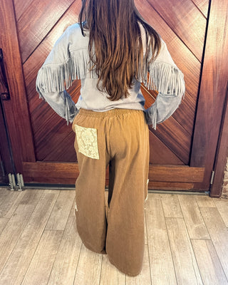 A person shown from the back wearing a light gray fringed top and wide-leg brown pants with a cream patch pocket, standing in front of a wooden door and wood floor.