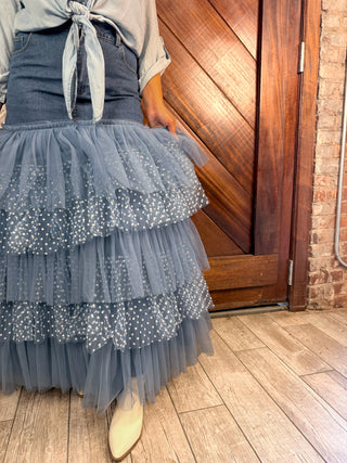 A person wearing a tiered tulle skirt in dusty blue-gray with white polka dots, paired with a denim apron-style top and beige ankle boots, standing indoors in front of a wooden door and brick wall.