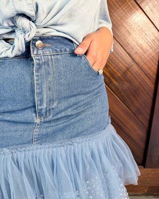 Close-up of a person wearing a light blue denim skirt with a ruffled tulle hem and a matching light blue top, standing against a dark wood panel background.