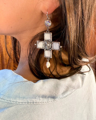 A close-up side profile of a woman wearing a large silver-toned cross dangle earring with white square stones and a small teardrop pearl charm, set against a background of brown hair and a light denim shirt.