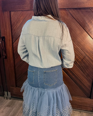 A person shown from the back wearing a light blue denim shirt and a blue denim skirt with a tulle overlay and small white embellishments, standing in front of a wooden door.