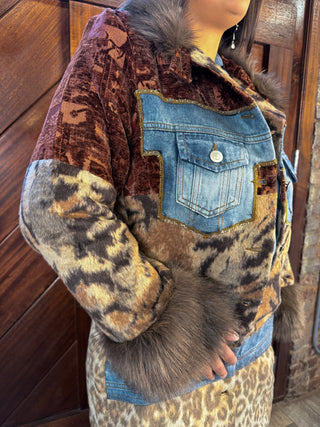 A close-up of a person wearing a patchwork-style jacket with a brown velvet chest panel, blue denim pocket, leopard-print sleeves and hem, and dark fur trim, standing in front of a wooden door.