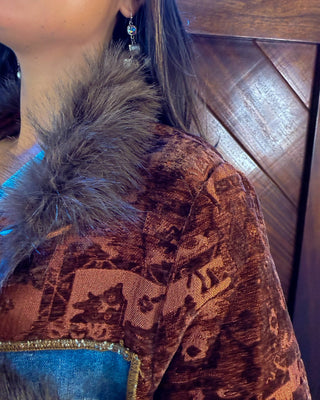 Close-up of a person wearing a brown patterned velvet top with a gray faux-fur collar and a blue fabric panel with gold trim, against a dark wood paneled background.