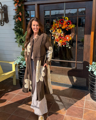 A woman stands on a tiled porch in front of a shop door decorated with an autumn leaf garland and a floral wreath. She is wearing a long brown cardigan with large cream floral patches over a striped outfit.