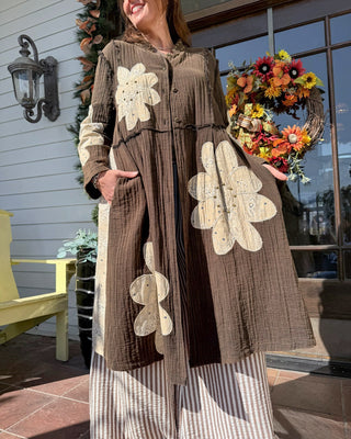 A woman wearing a brown textured longline cardigan with large cream-colored floral appliques, standing on a tiled porch in front of a house with a wreath and window.