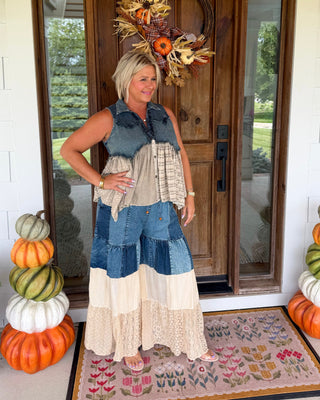 A woman stands on a front porch wearing a handmade patchwork maxi dress with denim blue and beige layers. The porch features a wooden door with a fall wreath, stacked pumpkins on both sides, and a patterned doormat.