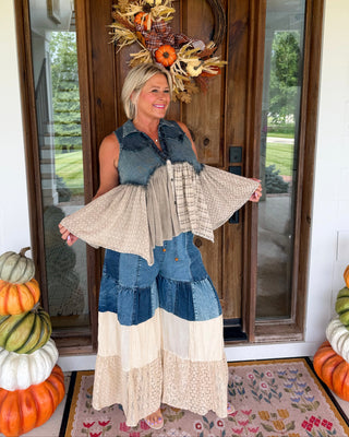 A woman stands on a front porch wearing a long patchwork dress with denim and cream sections, holding up a matching denim vest and cardigan. The porch has autumn decorations including stacked pumpkins and a fall wreath on the door.