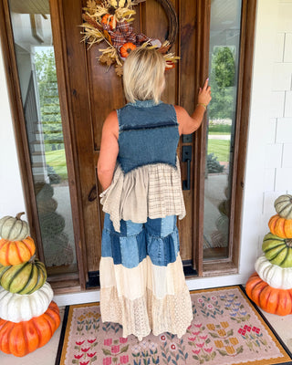 A woman stands with her back to the camera on a front porch, wearing a tiered patchwork dress made from blue denim and beige/cream fabrics. She is touching a fall wreath on a wooden door, with stacked pumpkins on both sides and a floral doormat in front.