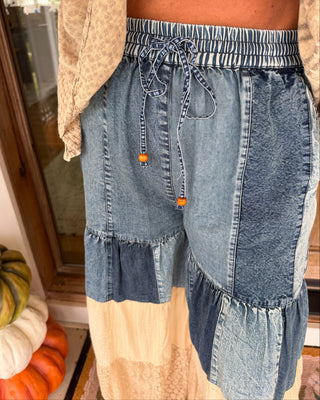A person wearing patchwork denim pants with a smocked elastic waistband, drawstring, and layered ruffle panels, paired with a cream shirt and skirt. Pumpkins are visible in the background near a window.