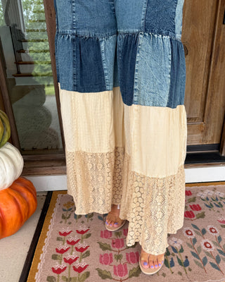 A person wearing a patchwork denim and cream pant with lace panels, standing on a decorative doormat at a front porch.