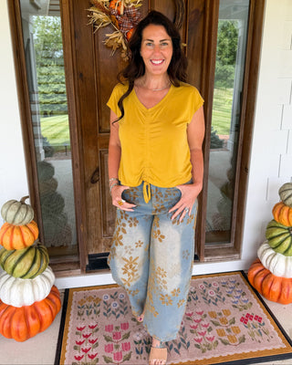 A woman stands on a front porch wearing a mustard yellow ruched short-sleeve top and light blue floral wide-leg pants. The porch has a wooden door with a fall wreath, stacked pumpkins on both sides, and a patterned doormat.