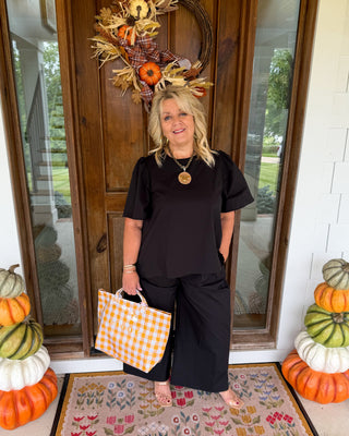 A woman stands on a front porch wearing a black short-sleeve top and matching wide-leg pants, holding a white and orange gingham tote bag. The porch has autumn decorations including a wreath on the door, stacked pumpkins on both sides, and a floral doormat.