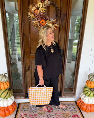A woman stands on a front porch holding a yellow and white gingham check tote bag. She is wearing a black short-sleeve top and black pants, standing on a floral doormat in front of a wooden door decorated with a fall wreath.