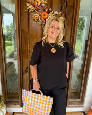 A woman stands on a front porch wearing a black short-sleeve top and black pants, holding an orange and white gingham tote bag with 'SOLD' printed on the front. She is posed in front of a wooden door decorated with a fall wreath.
