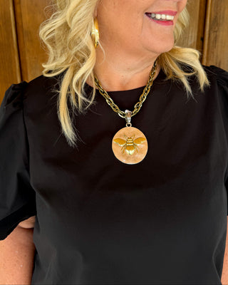 A smiling blonde woman wearing a gold chain necklace with a round peach pendant featuring a gold bumblebee, paired with a black top and gold hoop earrings, set against a wooden background.