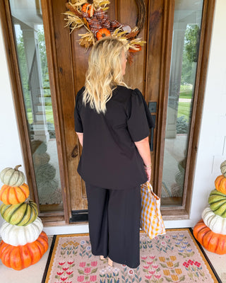 A woman stands with her back to the camera on a front porch, wearing a black short-sleeve top and matching wide-leg pants, holding a yellow-and-white gingham tote bag. The porch has stacked pumpkins on both sides, a floral doormat, and a decorative autumn wreath on the wooden door.