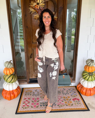 A woman stands on a front porch wearing a cream short-sleeve tie-front top and brown wide-leg pants with white floral patches. She is holding a teal handbag and standing on a patterned doormat in front of a wooden door decorated with autumn wreath and stacked pumpkins.