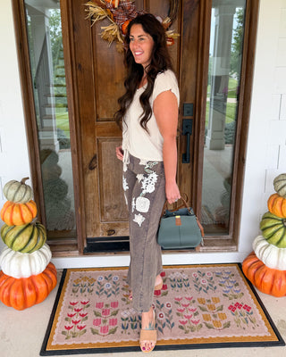 A woman stands on a front porch wearing a cream sleeveless top and brown patterned wide-leg pants, holding a teal handbag. The porch has a wooden door with a fall wreath, a patterned doormat, and stacked pumpkins on both sides.
