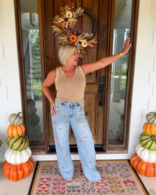 A woman stands on a front porch wearing a beige tank top and light blue high-waisted flare jeans with embroidered floral details. She is posing in front of a wooden door with a fall wreath, with stacked pumpkins on both sides and a patterned doormat beneath her.