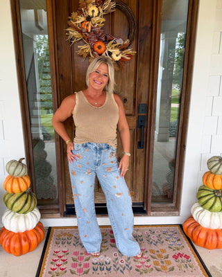 A woman stands on a front porch wearing a beige tank top and light blue high-waisted wide-leg jeans with small embroidered floral details. She is posing in front of a wooden door decorated with an autumn wreath, with stacked pumpkins on both sides and a floral doormat underneath.