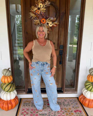 A woman stands on a front porch wearing a beige tank top and light blue distressed wide-leg jeans, with a fall wreath on the door behind her. Decorative stacked pumpkins sit on both sides of a floral doormat.
