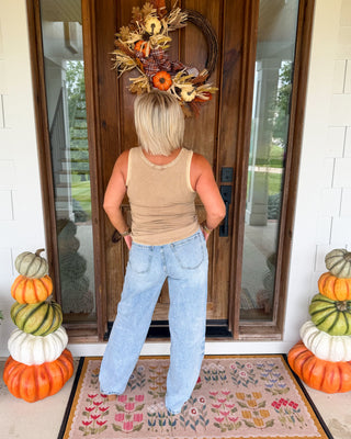 A woman stands with her back to the camera on a front porch, wearing a beige tank top and light blue jeans. She is positioned in front of a wooden door decorated with a fall wreath, with stacked pumpkins on both sides and a patterned doormat beneath her.