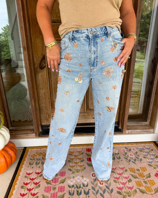 A person wearing light-wash blue jeans with scattered metallic rose-gold and gold floral appliqués, standing on a patterned doormat in front of a glass door.