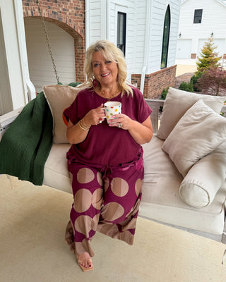A woman sitting on a white porch swing holding a mug, wearing a matching burgundy and beige polka-dot lounge set in front of a white house.