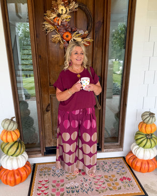 A woman stands on a front porch wearing a burgundy short-sleeve top and a magenta and beige patterned maxi skirt, holding a mug. The porch has a wooden door with a fall wreath, stacked pumpkins on both sides, and a floral doormat.