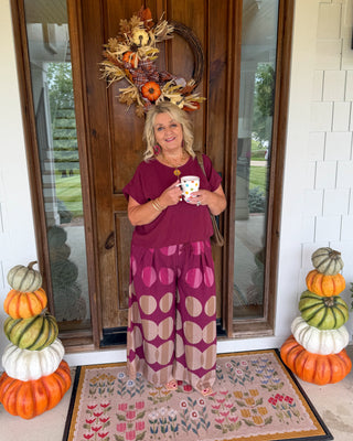 A woman stands on a front porch wearing a burgundy tunic top and a matching long skirt with large beige and pink oval patterns. She holds a white mug with a floral print.