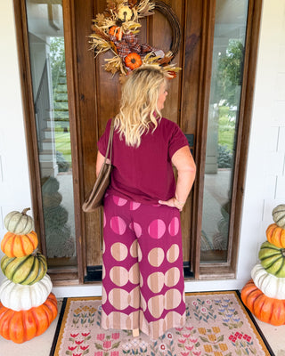 A woman stands with her back to the camera in front of a wooden front door, wearing a burgundy t-shirt and a matching wide-leg skirt with large beige and pink polka dots. She has a fall wreath on her head and is posing on a floral doormat with stacked pumpkins on either side.