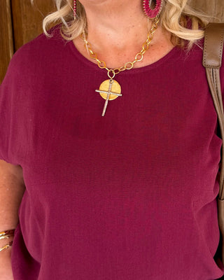 A woman wearing a burgundy short-sleeve top and matching patterned pants with pink and purple abstract shapes, standing in front of a wooden door with a fall wreath.