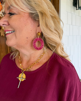 A woman wearing a gold chain necklace with a silver cross pendant and a large pink and gold spiral drop earring, shown in a close-up profile shot.