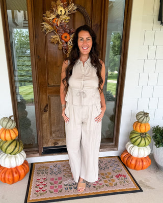 A woman stands on a front porch wearing a sleeveless light beige wide-leg jumpsuit. The porch has a wooden door with a fall wreath, stacked pumpkins on both sides, and a patterned doormat.