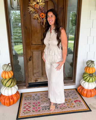 A woman stands on a front porch wearing a sleeveless cream-colored ruffled jumpsuit. The porch features a wooden door with an autumn wreath, stacked pumpkins on both sides, and a patterned doormat.