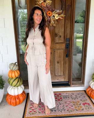 A woman stands on a front porch wearing a sleeveless light beige wide-leg jumpsuit. The porch has a wooden door with a fall wreath, stacked pumpkins on both sides, and a patterned doormat.