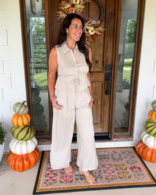 A woman stands on a front porch wearing a sleeveless, light beige wide-leg jumpsuit. The porch has a wooden door with a fall wreath, stacked pumpkins on both sides, and a patterned doormat.