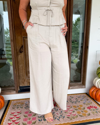 A woman models a light beige linen two-piece set consisting of a sleeveless peplum top and matching wide-leg pants, standing on a patterned doormat in front of a wooden door.