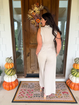 A woman stands with her back to the camera on a front porch, wearing a sleeveless cream-colored wide-leg jumpsuit. The porch features a rustic wooden door decorated with a fall wreath, a patterned doormat, and stacked pumpkins on both sides.