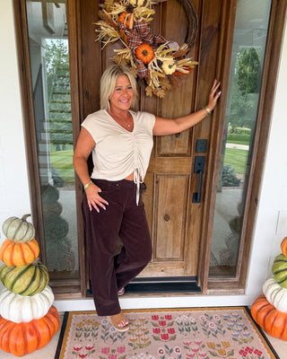 A woman stands on a front porch wearing a cream ruched short-sleeve top and dark brown wide-leg pants, with an autumn wreath on the door and stacked pumpkins on both sides.