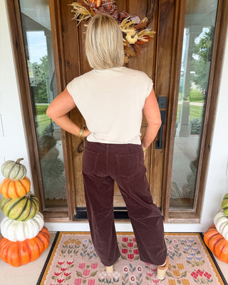 A woman stands with her back to the camera on a front porch, wearing a cream short-sleeve top and dark brown wide-leg corduroy pants. She is positioned in front of a wooden door decorated with a fall wreath, with stacked pumpkins on both sides and a patterned doormat beneath her.